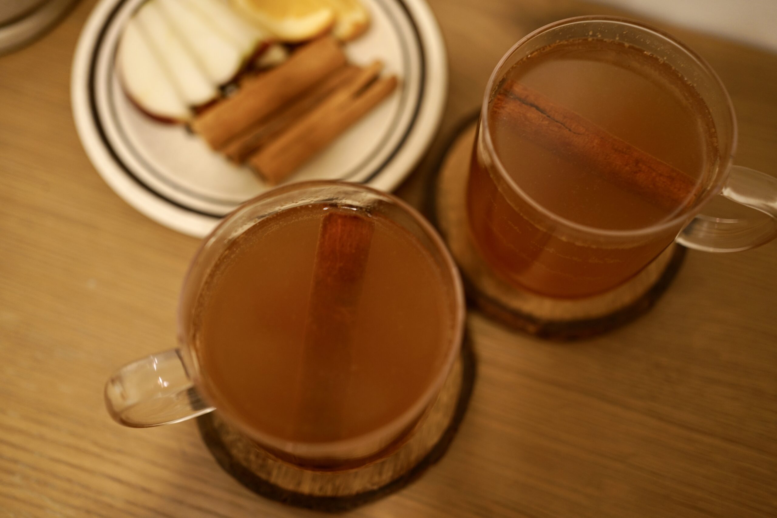 two mugs of apple cider with cinnamon sticks alongside a plate of toppings