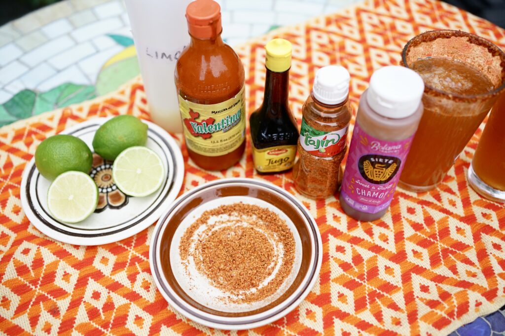 Micheladas next to limes, tajin, hot sauce, Maggi seasoning, and chamoy on a table