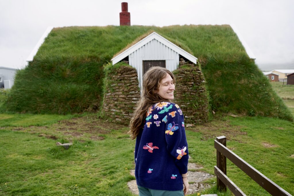 A woman poses in front of an Icelandic turf house